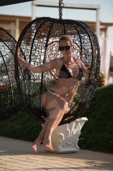 Young Woman Sitting in a Metal Rattan Hanging Basket With Cushions - Enjoying the Sunny Day in a Swimsuit wearing Sunglasses by the Sea