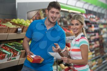 Beautiful Young Couple Looking At The Grocery List Shopping For Fruits And Vegetables In Produce Department Of A Grocery Store - Supermarket - Shallow Deep Of Field