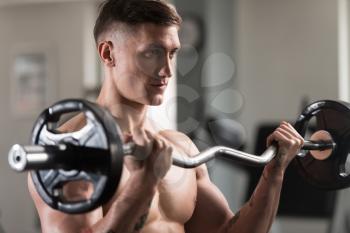 Young Muscular Man Doing Heavy Weight Exercise For Biceps With Barbell In Gym