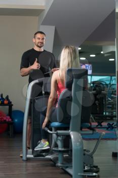 Personal Trainer Showing Ok Sign To Client - Young Woman Exercise Legs On Machine In The Gym Or Fitness Club