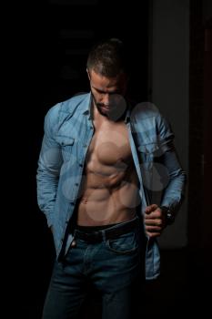 Healthy Young Man Standing Strong In The Gym And Flexing Muscles While Wearing Blue Shirt - Muscular Athletic Bodybuilder Fitness Model Posing After Exercises On Wall of Bricks