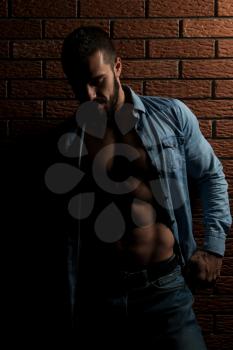 Healthy Young Man Standing Strong In The Gym And Flexing Muscles While Wearing Blue Shirt - Muscular Athletic Bodybuilder Fitness Model Posing After Exercises On Wall of Bricks