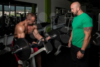 Mature Man Working Out In Gym - Doing Biceps Exercise On Machine With Help Of His Trainer
