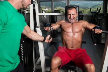 Mature Man Working Out In Gym - Doing Chest Exercise On Machine With Help Of His Trainer