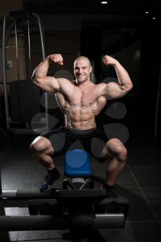 Healthy Young Man Sitting Strong In The Gym And Flexing Muscles - Muscular Athletic Bodybuilder Fitness Model Posing After Exercises