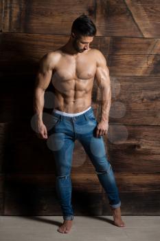 Healthy Young Man Standing Strong Against a Wooden Wall and Flexing Muscles While Wearing Blue Jeans - Muscular Athletic Bodybuilder Fitness Model Posing After Exercises - a Place for Your Text