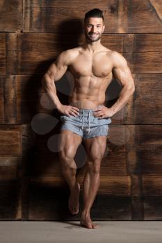Healthy Young Man Standing Strong Against a Wooden Wall and Flexing Muscles While Wearing Sports Shorts - Muscular Athletic Bodybuilder Fitness Model Posing After Exercises - a Place for Your Text