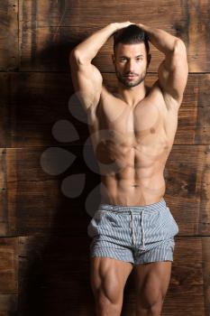Healthy Young Man Standing Strong Against a Wooden Wall and Flexing Muscles While Wearing Sports Shorts - Muscular Athletic Bodybuilder Fitness Model Posing After Exercises - a Place for Your Text