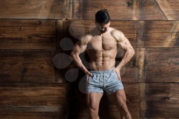 Portrait of a Young Physically Fit Man Showing His Well Trained Body While Wearing Sports Shorts - Muscular Athletic Bodybuilder Fitness Model Posing After Exercises on Wooden Wall - a Place for Your Text