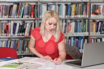 Pretty Woman With Blonde Hair Sitting at a Desk in the Library - Laptop and Organiser on the Table - Looking at the Screen a Concept of Studying - Blurred Books at the Back