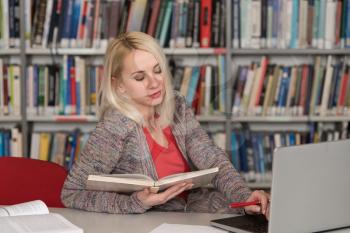 Pretty Woman With Blonde Hair Sitting at a Desk in the Library - Laptop and Organiser on the Table - Looking at the Screen a Concept of Studying - Blurred Books at the Back