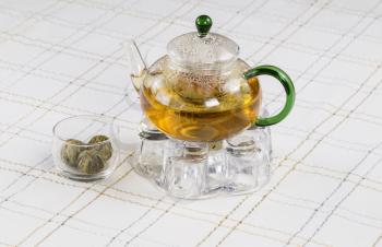 Horizontal photo of glass tea pot, holder and tea balls with White Striped Table Cloth in background