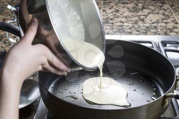 Horizontal photo of focus on pancake batter coming out of stainless steel bowl and into hot frying pan