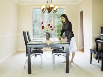Photo of mature woman placing diner mats in family formal dining room table with daylight coming through large windows in background