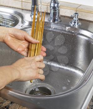 Vertical photo of female hands rinsing off bamboo chopsticks with running water from kitchen sink faucet in background
