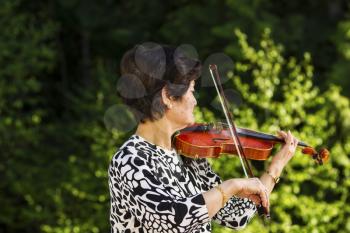 Horizontal photo of Senior Asian woman facing the forest while playing the violin outdoors with green trees in background