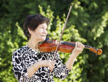 Horizontal photo of Senior Asian woman playing violin outdoors with bright green trees in background