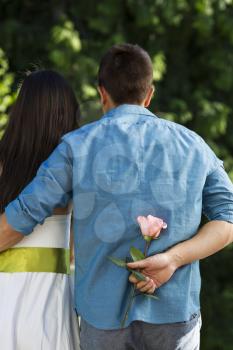 Vertical photo of a young couple facing away from the camera with man holding flower behind his back as surprised to woman with green trees in background