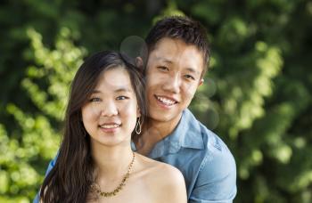 Horizontal photo of a young couple holding each other while looking forward with green trees in background