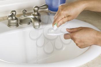 Horizontal Photo of female hands putting tooth paste on toothbrush with white bathroom sink, blue cup and running faucet in background 
