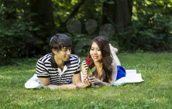 Horizontal photo of young adult couple, lying on blanket, while looking at single red rose with green grass and trees in background   