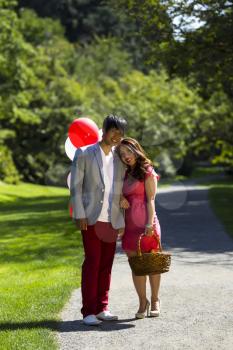 Vertical photo of young adult couple dressed in formal attire holding each other outdoors with several balloons, picnic basket, trail and trees behind them  