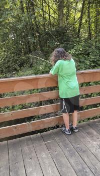 Vertical photo of young girl fishing off of wooden bridge for trout with stream and trees in background  
