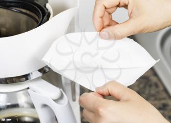 Horizontal photo of female hand opening up white bleached coffee filter with coffee maker, stone kitchen counter top and sink in background 