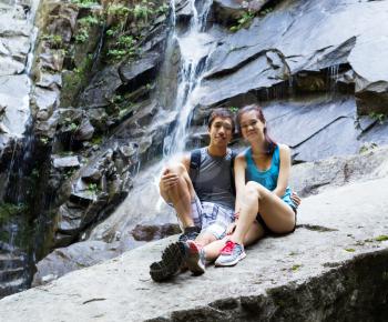 Photo of young adult couple resting near small waterfalls during their hike outdoors 