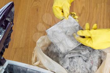Horizontal photo of gloved hands removing dirt from vacuum cleaner filter into plastic bag with hardwood floors in background 