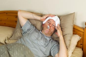 Horizontal photo of mature man holding wash cloth to his forehead along with thermometer in mouth while lying in bed 