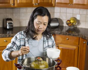 Photo of mature woman cooking winter melon from steaming pot with large spoon in hand and kitchen red oak cabinets in background 