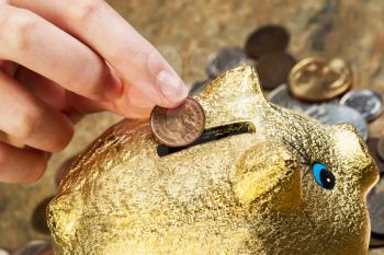 Horizontal photo of female fingers putting single cent into piggy bank with coins lying around on stone counter in background
