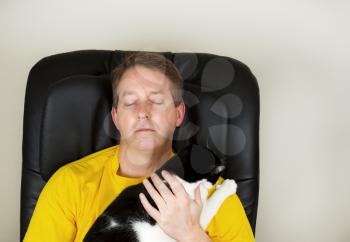 Photo of mature man relaxing in massage, with eyes closed, chair while holding a black and white cat