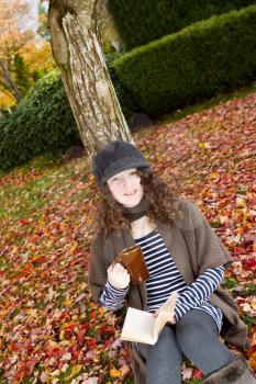Vertical photo of teenage girl, looking forward, enjoying her book, and drink, while lying against a tree during the autumn season dressed in earth colors  