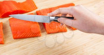 Horizontal photo of female hand holding sharp cutting knife with fresh pieces of Wild Red Salmon and natural bamboo board in background 