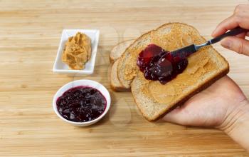 Horizontal photo of female hands making peanut butter and jelly sandwich with natural bamboo cutting board underneath