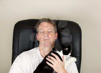 Photo of mature man wearing white shirt while relaxing in massage chair, eyes closed, with hands holding black and white family cat  
