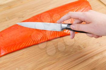 Horizontal photo of female hand holding sharp cutting knife with fresh Wild Red Salmon and natural bamboo board in background 