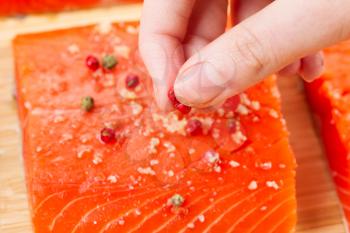 Horizontal photo of female fingers placing red peppercorns on fresh pieces of Wild Red Salmon and natural bamboo board in background 