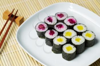 Horizontal photo, focus in upper part of plate, of pickled hand rolled sushi on white plate with chop sticks and holder in background and natural unstained bamboo dinner mat underneath