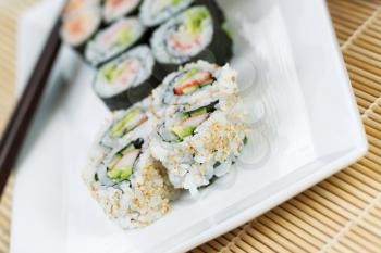 Closeup horizontal angled photo of freshly handmade California sushi rolls in white plate and chop sticks in background with bamboo mat underneath 