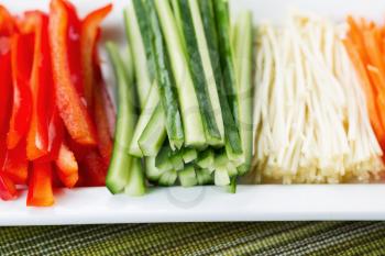 Horizontal closeup photo of freshly sliced cucumbers, placed in white plate, along with sliced cuts of carrots, red peppers and golden needle mushrooms with textured table cloth underneath