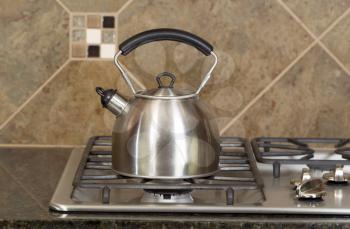 Horizontal photo of a stainless steel tea pot on stove top with stone counter tops and tile back splash 