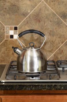 Vertical photo of a stainless steel tea pot on stove top with stone counter tops and tile back splash 