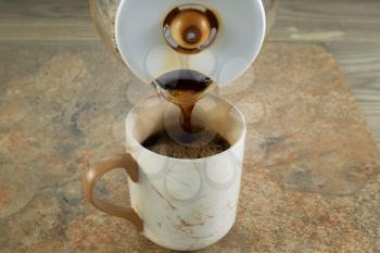 Horizontal photo of freshly brewed black coffee being poured from pot into a ceramic cup of with stone and wood underneath