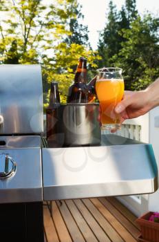 Vertical photo of male hand holding glass of beer with bucket of beer bottles, barbecue cooker and trees with daylight coming through in background 