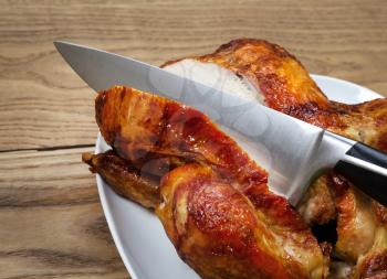 Side view of a freshly oven roasted whole chicken being sliced by large kitchen knife in white serving dish placed on rustic wood