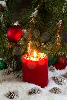 Vertical front view of a glowing red candle, resting in white snow, with evergreen tree branch and rustic wood in background 