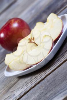 Angled vertical photo of fresh apple slices, on white plate, with whole apple and rustic wood in background  